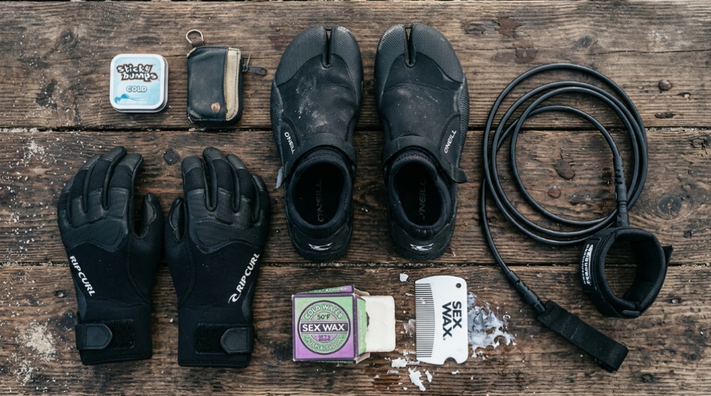 Cold-water surfing accessories including booties, gloves, and wax on a wooden board.